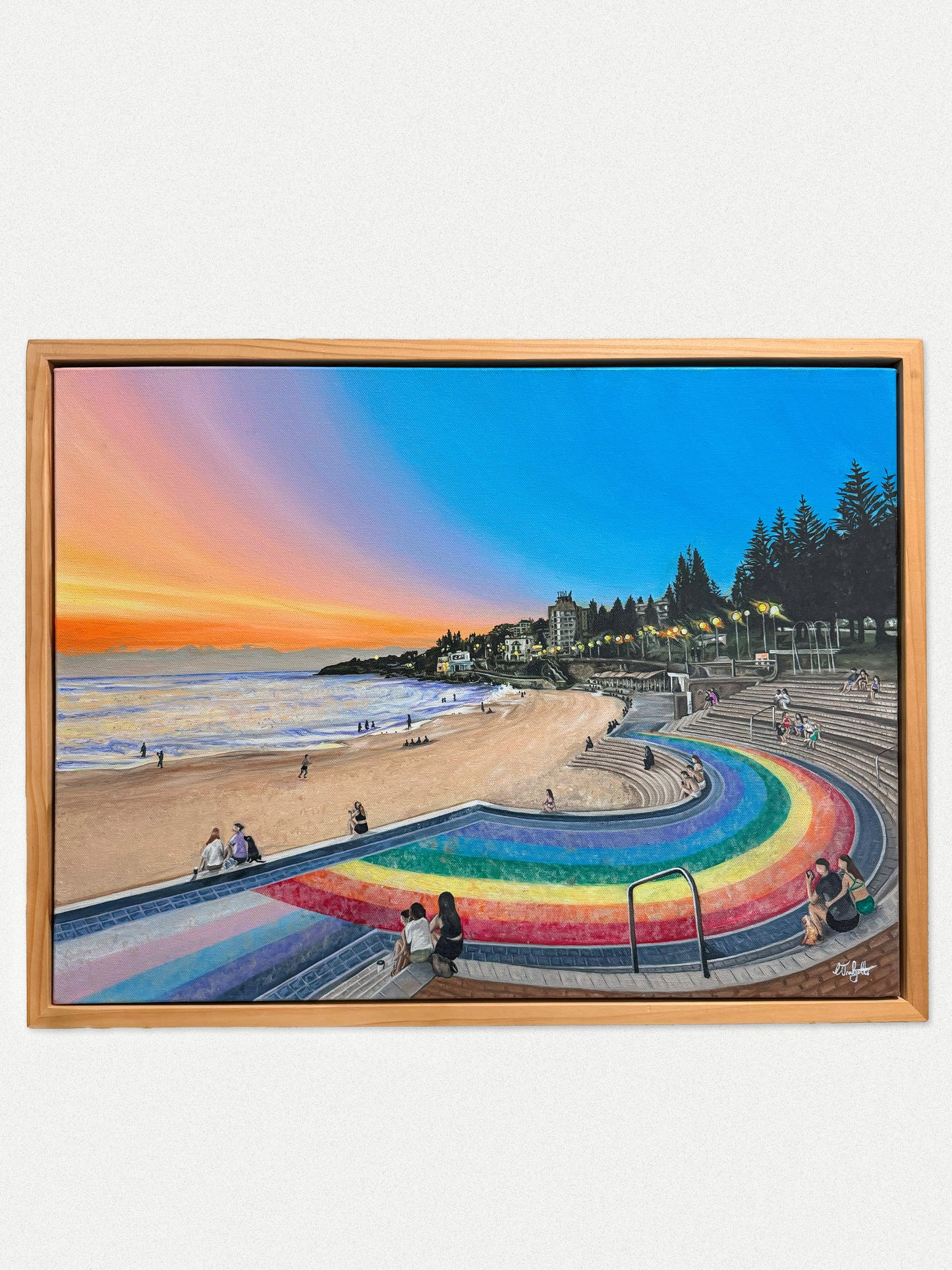 A framed painting of Coogee beach and  its iconic rainbow steps at sunrise, featuring a rainbow-colored curved walkway, ocean waves, sandy shore, trees, and people sitting along the steps. The artwork is housed in a light natural wood frame and displayed against a plain off-white wall.

